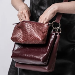 Pode incluir: Um conjunto de bolsas bordô de duas peças. A bolsa superior é uma pequena bolsa texturizada com zíper. A bolsa inferior é uma bolsa retangular maior e brilhante. Ambas as bolsas são conectadas por uma alça bordô com anéis prateados.