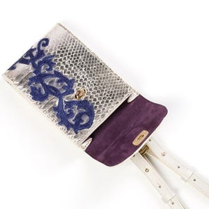 Puede incluir: Un bolso de mano plateado metalizado con textura de piel de serpiente, con un patr&oacute;n floral bordado en azul oscuro en la solapa frontal. El interior est&aacute; forrado con tela morada oscura e incluye correas blancas finas ajustables con herrajes.