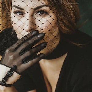 May include: A portrait featuring a person wearing a black veil and sheer gloves. The gloves have a leather strap with a buckle. The person is wearing a black choker and a black top. The background is blurred.