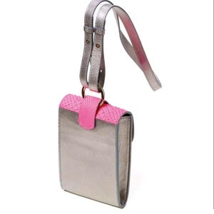Puede incluir: Un bolso bandolera de color plata metalizado con una solapa de color rosa intenso texturizada con un patr&oacute;n similar a la piel de serpiente. El bolso incluye una correa ajustable a juego fijada a una anilla met&aacute;lica circular en la parte superior.