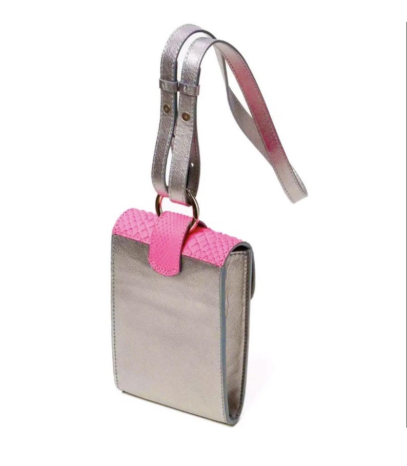 Puede incluir: Un bolso bandolera de color plata metalizado con una solapa de color rosa intenso texturizada con un patr&oacute;n similar a la piel de serpiente. El bolso incluye una correa ajustable a juego fijada a una anilla met&aacute;lica circular en la parte superior.