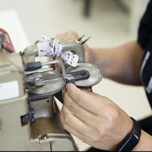 Puede incluir: Una m&aacute;quina de coser industrial de color beige con un asa met&aacute;lica envuelta en una tira de tela estampada. La m&aacute;quina cuenta con dos placas met&aacute;licas circulares y un mecanismo de aguja para coser, con un dial de control negro en el lateral.
