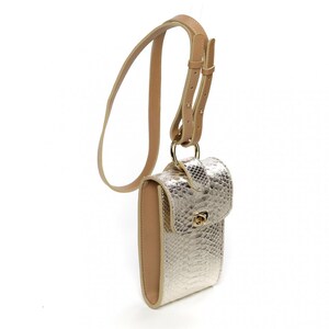 Puede incluir: Un bolso bandolera rectangular vertical con un acabado texturizado de piel de serpiente en color dorado met&aacute;lico. El bolso incluye un borde color cuero beige, un cierre giratorio de metal dorado en la solapa y una correa ajustable a juego.