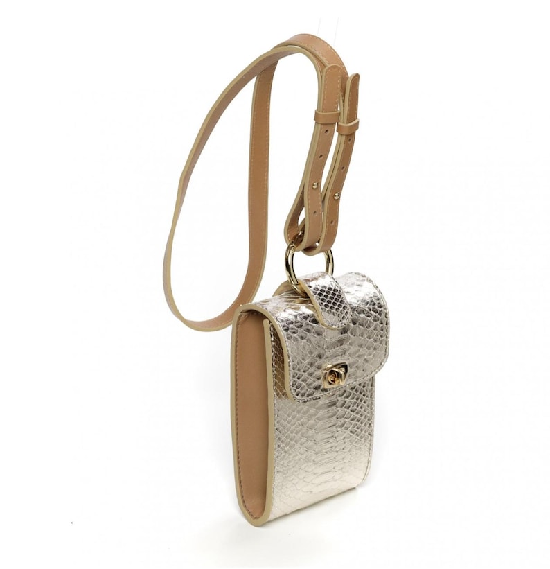 Puede incluir: Un bolso bandolera rectangular vertical con un acabado texturizado de piel de serpiente en color dorado met&aacute;lico. El bolso incluye un borde color cuero beige, un cierre giratorio de metal dorado en la solapa y una correa ajustable a juego.