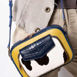 Peut inclure: Sac bandoulière au design unique. Le sac est composé d'un corps en cuir jaune avec une bordure et une bandoulière bleues. L'avant présente un panneau en peau de vache blanche et noire et un rabat bleu marine. Le sac a une fermeture éclair dorée.