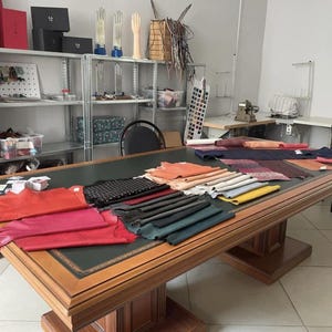 Puede incluir: Una mesa grande de madera con una superficie de cuero verde oscuro cubierta con varios montones de muestras de cuero de colores en tonos rojo, rosa, negro, azul y amarillo, situada en un taller con estanter&iacute;as met&aacute;licas con suministros.