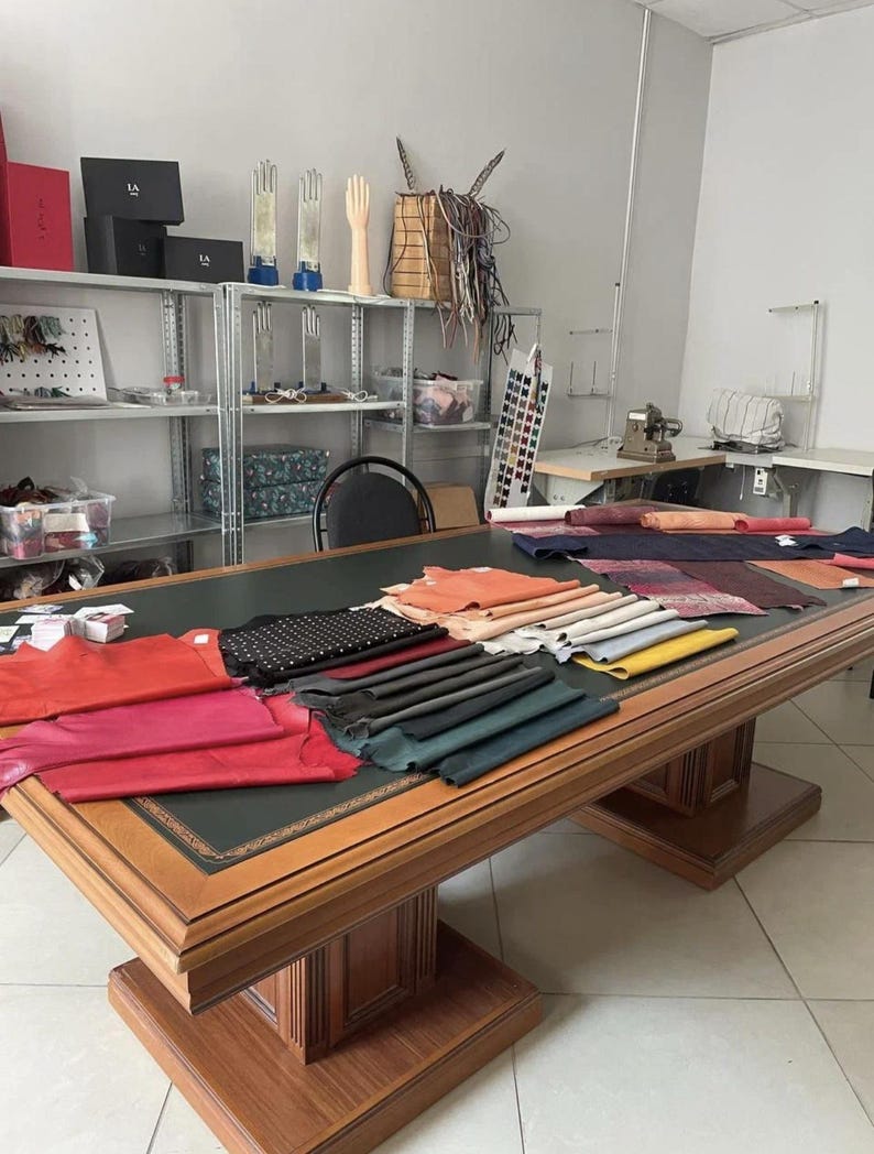 Puede incluir: Una mesa grande de madera con una superficie de cuero verde oscuro cubierta con varios montones de muestras de cuero de colores en tonos rojo, rosa, negro, azul y amarillo, situada en un taller con estanter&iacute;as met&aacute;licas con suministros.