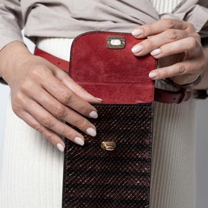 Pode incluir: Uma bolsa de cinto retangular vertical com um exterior de textura de pele de cobra castanho-escuro e um forro interior de camurça vermelho profundo. A bolsa inclui um fecho de metal dourado na aba frontal e uma correia de cinto em pele vermelha.