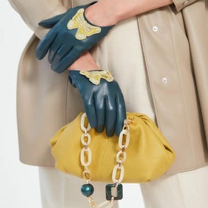 Könnte beinhalten: Teal-farbene Lederhandschuhe mit goldfarbenem Schmetterlingsapplikation. Eine senfgelbe Clutch mit Kettenriemen wird von den Handschuhen gehalten. Der Kettenriemen hat weiße und goldfarbene Glieder mit einem dunkelgrünen Perlen- und Quadratakzent.