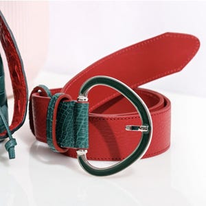 Puede incluir: Un cinturón de cuero rojo con superficie texturizada y una trabilla de cuero verde con relieve de cocodrilo. El cinturón cuenta con una hebilla grande de metal plateado de forma redondeada con una incrustación de esmalte verde.