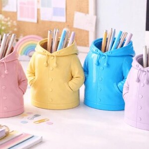 May include: Four colourful pen holders shaped like hooded jackets. The pen holders are in pastel shades of pink, yellow, blue, and lavender. Each holder is filled with pens and pencils. The design includes a hood, buttons, and pockets.