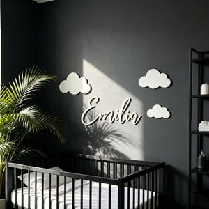 Può includere: Una scritta da parete bianca che riporta il nome Emilia in carattere corsivo, accompagnata da tre decorazioni murali a forma di nuvola bianca. Gli elementi sono fissati su una parete grigio scuro sopra una culla nera con biancheria bianca.