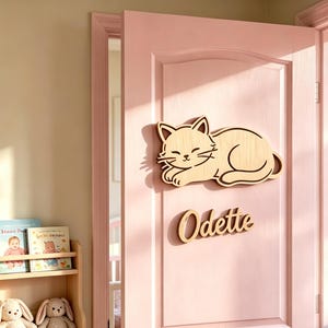 以下が含まれることがあります： ピンク色のドアに取り付けられた、猫の形をした木製の壁飾り。猫は目を閉じて横たわっています。猫の下には筆記体で「Odette」という名前があります。背景には、子供向けの絵本とぬいぐるみが入った木製の本棚があります。