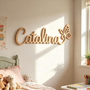 Puede incluir: Letrero de nombre de madera con el nombre "Catalina" y un diseño de mariposa. Las letras en cursiva son de color marrón claro, montadas en una pared blanca. Es un elemento decorativo para un dormitorio infantil.