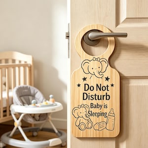 May include: A wooden door hanger with a light wood grain finish. The hanger features a black illustration of an elephant, stars, a baby bottle, and the text "Do Not Disturb Baby is Sleeping."