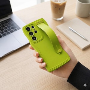 Puede incluir: Una funda de teléfono verde brillante con una correa a juego, sostenida en una mano. La funda tiene un diseño texturizado y un módulo de cámara con múltiples lentes. La palabra "fashion" está impresa en la parte superior de la funda.