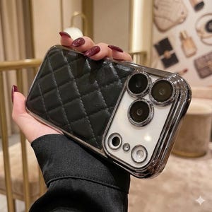 May include: Black quilted phone case with a silver frame. The case has a diamond pattern and a shiny silver border around the camera lenses. The phone case is designed to protect the phone from scratches and damage.