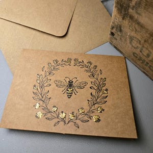 May include: A brown card with a black and gold bee illustration surrounded by a wreath of black leaves and gold flowers.