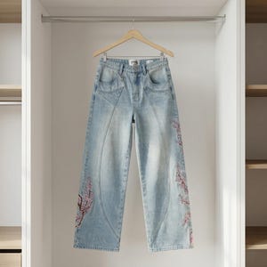 Op de afbeelding: Lichtblauwe jeans met geborduurde bloemmotieven in rood en roze. De jeans heeft een hoge taille en een wijde pijp. Het hangt aan een houten hanger in een witte kast.