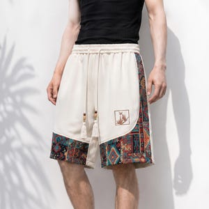 Op de afbeelding: Off-white shorts met een elastische tailleband en lange trekkoorden die eindigen in houten kralen en kwastjes. De onderkant en zijpanelen zijn voorzien van een levendig, meerkleurig geometrisch en bloemmotief met een kleine vierkante patch.