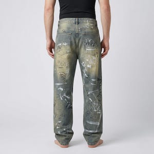 May include: Faded blue denim jeans with white graffiti-style designs. The jeans have a relaxed fit and a light wash, with distressed details. The back pockets are visible, and the jeans are worn by a person.