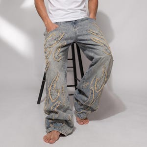 Peut inclure: Jean en denim bleu clair avec une coupe large et décontractée et un délavage usé. Le pantalon est orné de nombreuses applications en tissu en forme de flammes aux bords effilochés, créant un aspect texturé et superposé sur les jambes.