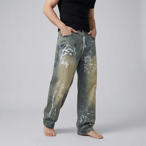 May include: Faded blue denim jeans with white paint-like designs. The jeans have a relaxed fit and a light wash, with distressed details. The overall style is casual and artistic.