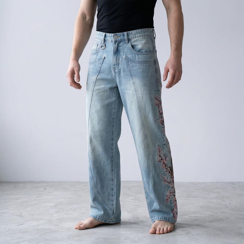 Op de afbeelding: Lichtblauwe denim jeans met een relaxte pasvorm. De jeans hebben een decoratieve bloemenborduurwerk in roze en rode tinten langs de zijkant van &eacute;&eacute;n been. De jeans hebben een uniek naadontwerp en een metalen ringdetail aan de voorkant.