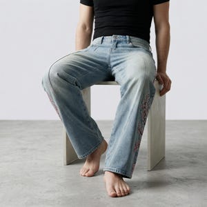 Op de afbeelding: Lichtblauwe denim jeans met decoratieve bloemenborduursels langs de zijnaden. De jeans hebben een relaxte pasvorm en een klassiek vijf-zakken ontwerp. Het model draagt een zwart t-shirt.