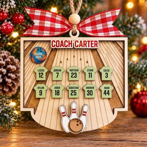 May include: A wooden bowling-themed Christmas ornament featuring the text COACH CARTER, a blue and orange bowling ball with TIGERS 2026, and ten light green jersey-shaped plaques with names and numbers, topped with a red and white gingham bow.