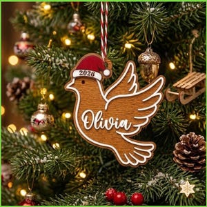 Peut inclure: Une décoration en bois en forme de colombe portant un bonnet de Père Noël rouge avec l'inscription 2026 et le prénom Olivia en écriture blanche. L'oiseau est suspendu par une ficelle rayée rouge et blanche avec un contour blanc.