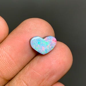 May include: A heart-shaped opal gemstone featuring a vibrant play-of-color with iridescent flashes of blue, green, and pink. The polished stone displays a smooth, rounded surface with a distinct heart silhouette, showcasing a bright, multi-colored pattern.