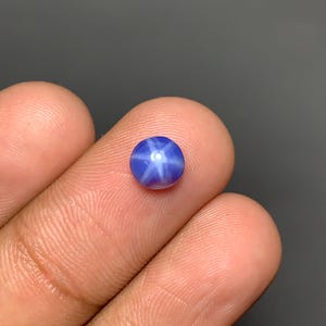 May include: A small, round, cabochon-cut blue gemstone featuring a distinct six-rayed star effect known as asterism. The stone displays a deep, saturated blue color with a bright, centered white star pattern that reflects light across its surface.