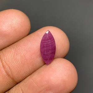 May include: A marquise-cut gemstone with a deep magenta and purple hue, featuring a faceted surface and visible internal striations. The stone has a pointed, elongated oval shape with a polished finish, displaying a distinct color gradient.