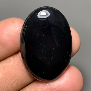 May include: A smooth, oval-shaped black gemstone. The stone has a shiny surface with a small, white, circular inclusion near the top. The gemstone is held in a person's hand, with the background out of focus.