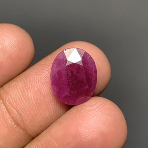 Op de afbeelding: Een ovaalvormige, gefacetteerde robijn met een diepe magenta-rode kleur. De steen heeft een gepolijst oppervlak met meerdere driehoekige en vliegervormige facetten die het licht reflecteren, wat een doorschijnende kwaliteit en een rijke, verzadigde tint laat zien.