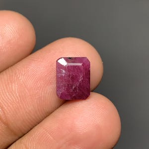 May include: A rectangular, emerald-cut ruby gemstone with a deep magenta-red hue. The stone features a faceted surface that reflects light, showing internal natural inclusions and a polished finish, held between two fingers against a dark background.