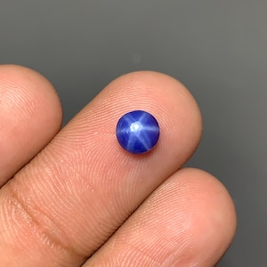 May include: A small, round, cabochon-cut blue star sapphire gemstone featuring a distinct six-rayed star pattern that appears on its surface. The stone displays a deep, saturated blue color and a smooth, polished finish, reflecting light clearly.