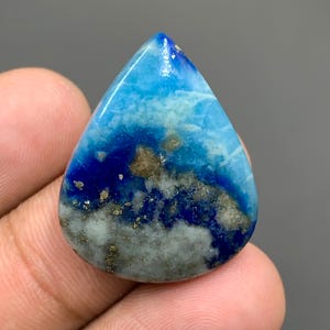 May include: A teardrop-shaped polished gemstone featuring a vibrant gradient of sky blue, deep royal blue, and white, with scattered metallic gold-colored pyrite inclusions throughout the surface, showcasing a smooth, reflective, and glossy finish.
