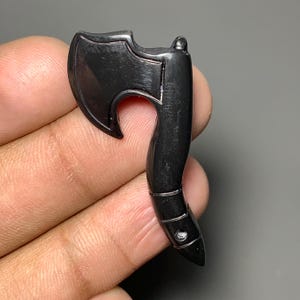 Può includere: Un piccolo ciondolo a forma di ascia nera con un manico curvo e una testa d'ascia dettagliata. Il ciondolo è realizzato in un materiale scuro e lucido con una finitura liscia. La testa dell'ascia ha una lama curva e un piccolo foro vicino al manico.