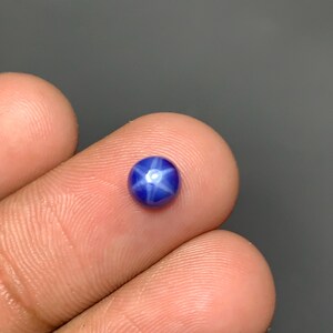 May include: A small, round, cabochon-cut blue gemstone featuring a distinct six-rayed star effect centered on its surface. The stone displays a deep, saturated blue color with a bright, reflective white star pattern that appears to float within the gem.