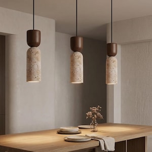 Puede incluir: Tres l&aacute;mparas colgantes con un cuerpo cil&iacute;ndrico de piedra beige texturizada y una parte superior de madera marr&oacute;n oscuro. Las luces cuelgan de cables negros, iluminando una mesa de madera. La escena sugiere un comedor moderno.