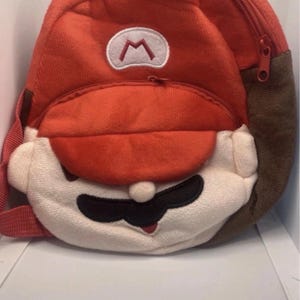 May include: A red plush backpack resembling a popular video game character. The backpack has a red body, a white face, and a black moustache. The character's hat has a white emblem. The backpack has a red zip and brown side panels.