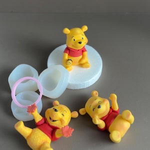 Könnte beinhalten: Eine Sammlung gelb-roter Winnie-the-Pooh-Figuren in verschiedenen Posen, darunter sitzend und liegend. Ebenfalls enthalten sind Silikonformen und ein rosa Ring, wahrscheinlich zum Basteln oder Backen. Der Hintergrund ist neutral.