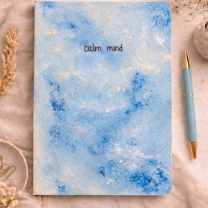 Può includere: Un diario blu con le parole "calm mind" in nero. La copertina presenta un design ad acquerello nei toni del blu e del bianco. Una penna azzurra con dettagli dorati è appoggiata accanto al diario.