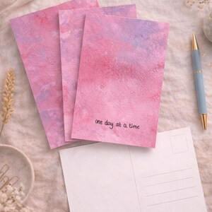 Könnte beinhalten: Drei rosa und lila Aquarellpostkarten mit dem Text "one day at a time" sind auf einer weißen Postkarte gestapelt. Ein blauer Stift mit goldener Spitze und dekorative Zweige sind ebenfalls im Bild.