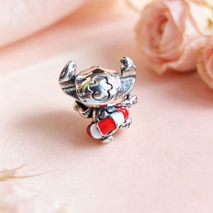 May include: A silver charm bead shaped like the character Stitch, featuring large ears and a wide head. The character is holding a small, round life preserver ring that is decorated with alternating horizontal bands of bright red and white enamel.