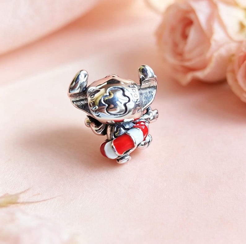 May include: A silver charm bead shaped like the character Stitch, featuring large ears and a wide head. The character is holding a small, round life preserver ring that is decorated with alternating horizontal bands of bright red and white enamel.