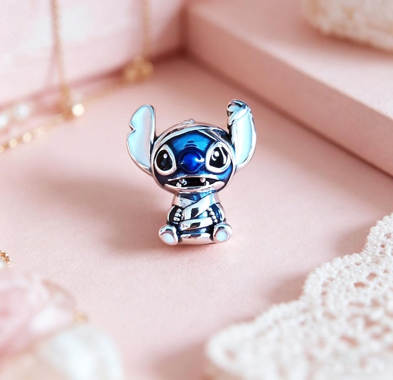 May include: A detailed, silver-toned charm featuring the character Stitch from the animated film Lilo & Stitch. The charm is primarily blue with white accents on the ears and feet. The charm is set against a soft pink background.