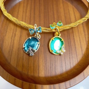 May include: Two gemstone pendants with gold and silver accents. One pendant is oval-shaped with a teal stone and a silver bee charm. The other is round with a green stone and a butterfly charm. Both have bow-shaped accents.